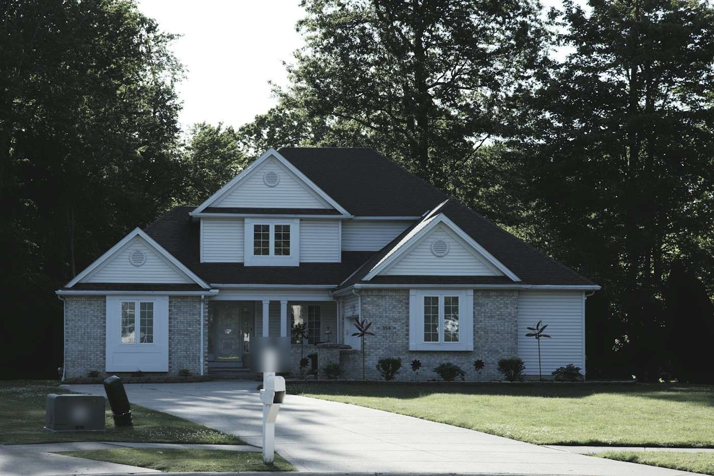 A gray two-story house with a dark shingle roof on a wooded lot
