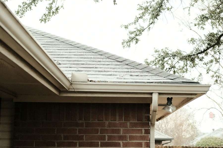 The shingle roof edge, gutter and downspout at the corner of a brick house